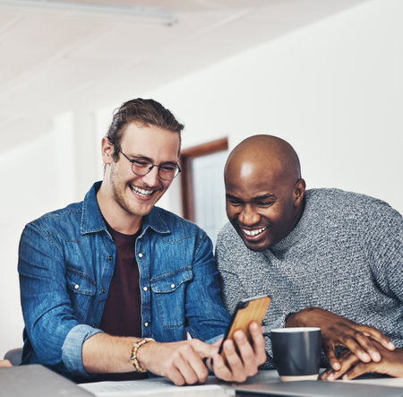 This is the funniest thing Ive seen all year. two cheerful young businessmen browsing on a cellphone together inside of the office during the day.の写真素材