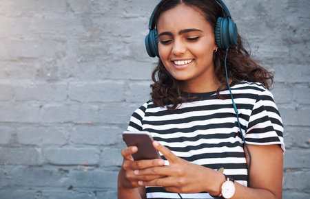 Music can get me through any day. a young woman using a cellphone while wearing headphones against a...