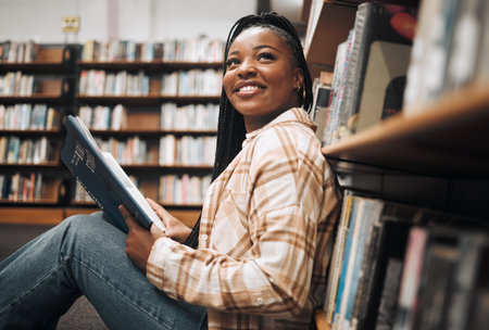 University, education and black woman in library reading book. College scholarship, student and happy female learner thinking about studying, knowledge and learning while sitting by bookshelf or caseの写真素材