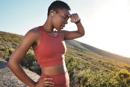 Fitness, music and tired with a sports black woman running in the mountain for cardio or endurance exercise. Workout, exhausted and sweat with a young female runner or athlete taking a breakの写真素材
