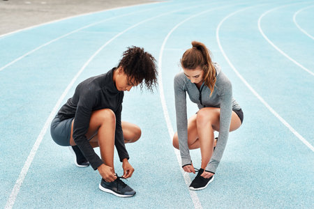 Laces tied, lets go. Full length shot of two attractive young athletes crouching together and tying their shoelaces before running on a track.の写真素材