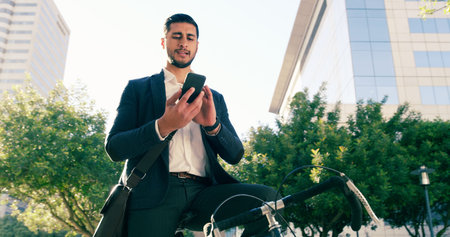 Sending a few message on his way to work. Low angle shot of a handsome young businessman using his cellphone while commuting to work on his bicycle.の写真素材
