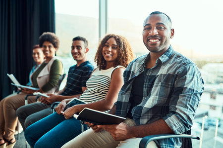 Portrait of a group of young cheerful work colleagues seated on chairs next to each other in a row...
