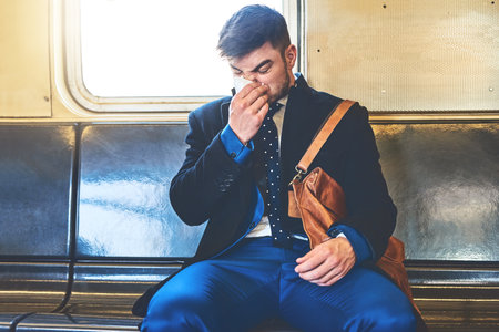 I better not be getting sick. a irritated looking young man blowing his nose while being seated inside of a train to get to work.の写真素材