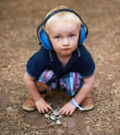 Keeping himself busy in nature. a little boy sitting and looking up at the camera with protective headphones over his ears.の写真素材