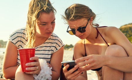 Texting more people to come to the beach. young female best friends hanging out at the beach.の写真素材
