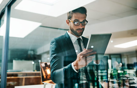 Looks like the company status is growing. a cheerful young businessman browsing on his digital tablet while standing inside of the office.の写真素材