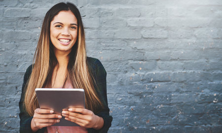 Who needs a cable to connect. Portrait of a young woman standing outdoors and using a digital tablet against a gray wall.の写真素材