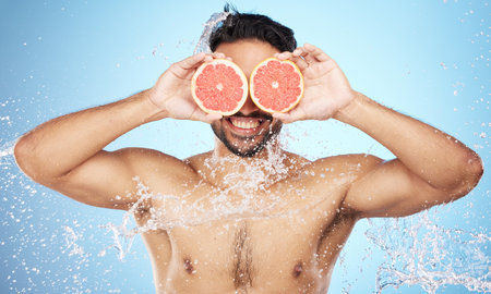Face, water and grapefruit with a man model in studio on a blue background for hygiene or natural hydration. Skincare, beauty or fruit with a handsome young male wet from a water splash in the showerの写真素材