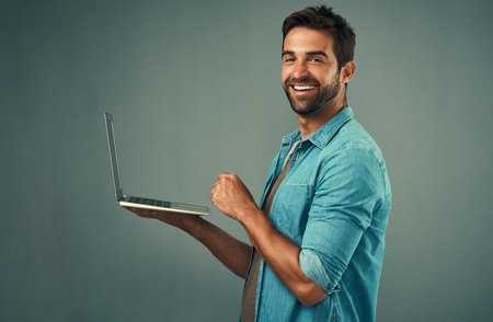 Hes never far from his tech. Studio portrait of a handsome young man using a laptop against a grey background.の写真素材