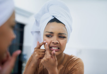 Pimple, begone. a young woman frowning while examining an imperfection on her skin in the mirror.の写真素材