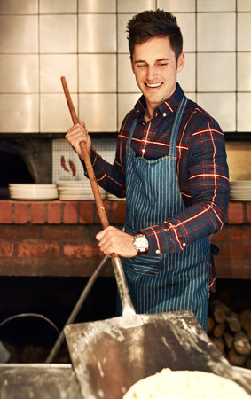 My pizza is what the customers come back for. a young man using a peel while making a pizza behind the counter of a pizzeria.の写真素材