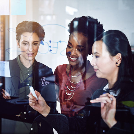 Mapping out a path to success. three businesswomen working on a glass wall the office.の写真素材