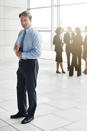 Confidence and success go hand-in-hand. Full length portrait of a businessman standing a lobby with his colleagues in the background.の写真素材