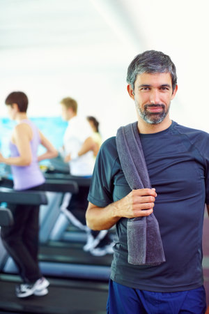 Man working out in a gym. Portrait of middle aged man holding towel with people exercising in background.の写真素材