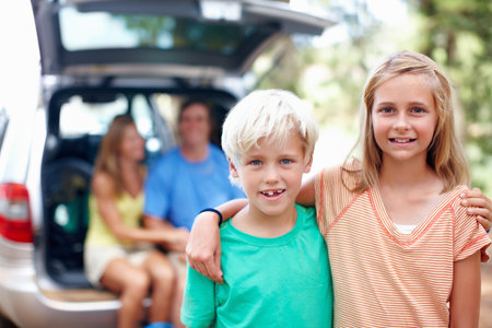 Brother and sister standing with arms around and smiling. Brother and sister standing with arms around and parents sitting in the back of a car.の写真素材