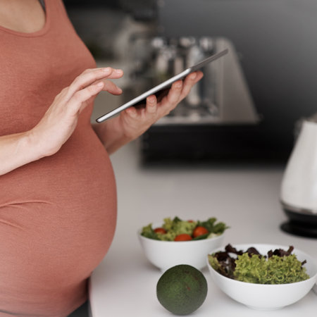 Checking her eating plan. a pregnant woman using her tablet while eating a salad in the kitchen.の写真素材