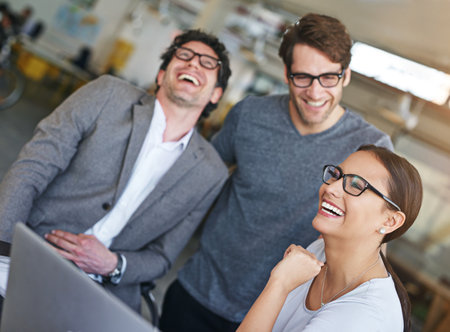 Success keeps them upbeat. laughing colleagues working together on a laptop in a modern office.の写真素材