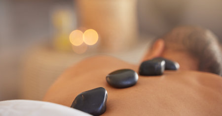 Spa, man and stone massage to relax, luxury and health for body care, peace and holistic care. Male, gentleman and lying on table at resort, rocks and wellness therapy for skincare and stress relief.の写真素材