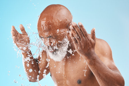 Black man cleaning face with water splash, healthy skincare on studio blue background for shower, cosmetics and body care transformation. Male model, senior beauty and facial hygiene with clean waterの写真素材