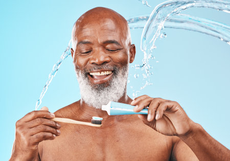 Water, splash and man with dental care in a studio for mouth health and wellness. Toothpaste, toothbrush and elderly African guy brushing his teeth for fresh oral hygiene isolated by blue background.の写真素材