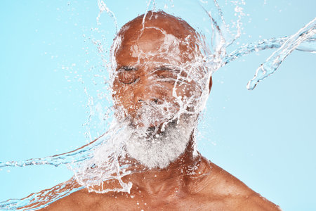 Elderly black man, water splash and face in studio for wellness, clean or facial health by backdrop. Man, model face and water for skincare, healthy self care or cosmetic beauty by blue backgroundの写真素材