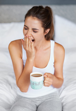 Still so sleepy...A young woman giving a big yawn while sitting in bed with a cup of coffee.の写真素材