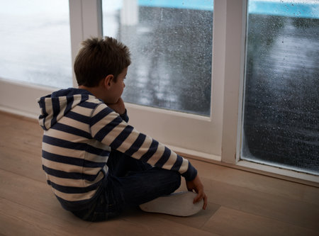 Rainy days with nothing to do...A young boy sitting by the window and looking bored while it rains outside.の写真素材
