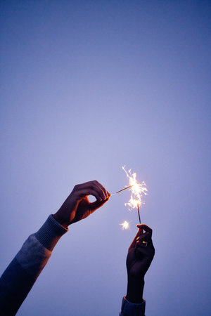 Sparkler, fire and hands of a person on a blue sky, celebration with light and outdoor party at...