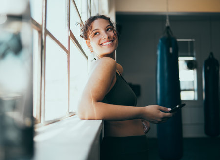 Window, smartphone and woman in gym, smile and online for training schedule, motivation and achievement. Happy, female athlete and girl with cellphone, break and connection for website and wellnessの写真素材