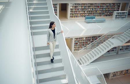 Woman, staircase and walking in the library for education, learning or knowledge from books in the...
