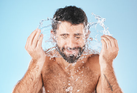 Face, splash and man in studio for cleaning, wellness and personal health with self care by blue background. Model, facial and healthy with water splash, skincare and cosmetics by studio backdropの写真素材