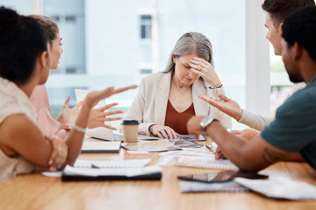 Group of businesspeople having a meeting together in a boardroom at work. Stressed mature caucasian businesswoman suffering from a headache while her colleagues talkの写真素材