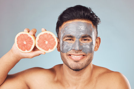 Portrait of one handsome young indian man applying a detoxifying clay or charcoal facial mask while holding grapefruit against a blue studio background. Guy using moisturising products with natural iの写真素材