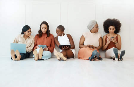 Group of five businesswomen sitting on the floor against a wall in an office and using tech. Businesspeople and colleagues talking and using technology while sitting in a row together at workの写真素材