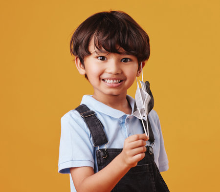 Portrait of an adorable little Asian boy happy while wearing a mask against an orange studio...