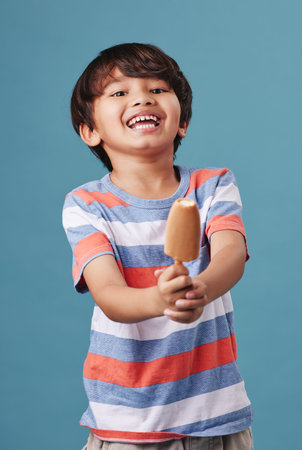 Portrait of an adorable little asian boy looking happy while enjoying a sweet treat against a blue...