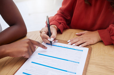 Closeup of two unknown ethnic business woman sitting and signing office contract. African american professional using hand gesture to point on paperwork. Mixed race colleague agreeing to document deaの写真素材