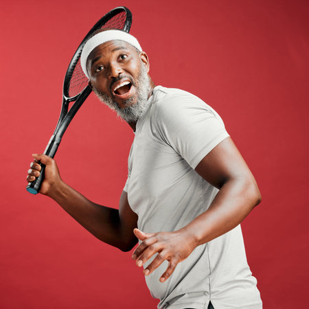 One happy mature african american man standing against a red background in studio and posing with a tennis racquet. Smiling black man feeling fit and sporty while playing a match. Ready for the courtの写真素材