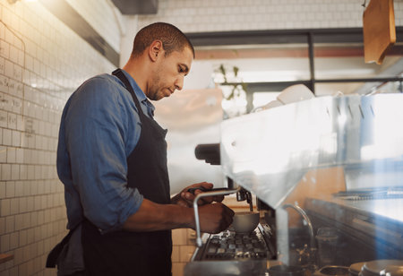 Focused barista making a cup of coffee. Barista making a cup of cappuccino. Coffeeshop assistant preparing a cup of espresso using the coffee machine. Mixed race businessman making a cup of coffeeの写真素材