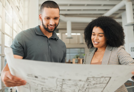 Young coworkers looking at a blueprint. Two businesspeople collaborate on a building plan. Design creatives working together in a modern office. Happy colleagues plan a building project togetherの写真素材