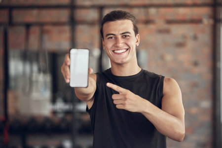 Happy man pointing to his phone in the gym. Portrait of a young man taking a break from exercise to use his cellphone. man online on his cellphone at the gym. Man gesturing toward his mobile phoneの写真素材