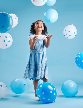 Adorable little girl looking up playing and having fun with blue and white balloons in celebration of her birthday against a blue studio backgroundの写真素材