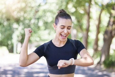 Cheerful young sporty female athlete celebrating while looking at smart watch. Hispanic sportswoman making winner gesture with clenched fist while tracking her progress while training outdoorsの写真素材