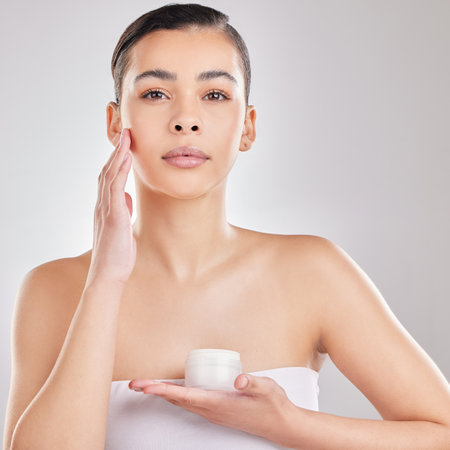 This product does wonders for my skin. a young woman applying a cream to her face against a grey background.の写真素材