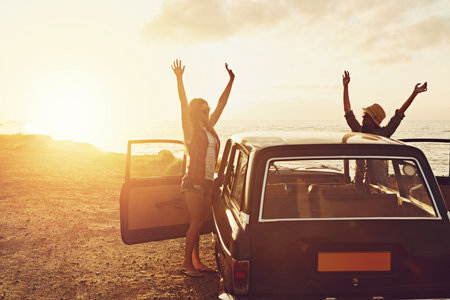 Sunset, road trip and couple of friends at beach for travel, journey and holiday, freedom and celebration. Celebrate summer and happy vintage or retro women with car for vacation and parking by oceanの写真素材