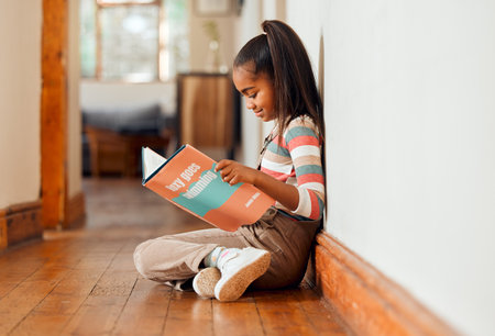 Little girl, book and reading on wooden floor for learning, education or story time relaxing at home with smile. Happy child smiling, sitting and enjoying books, read or stories at the house indoorsの写真素材