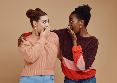 Gossip, friends and surprise face of young women with a secret and brown studio background. Wow, student surprised and girl chat together with whisper and excited discussion with shock from storyの写真素材