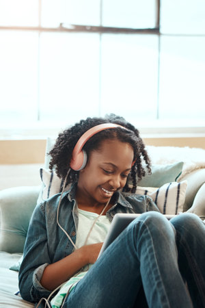 Digital tablet, sofa and girl with headphones relaxing while listening to music, radio or podcast. Rest, technology and African child watching a video or movie on mobile device in living room at homeの写真素材
