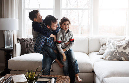 Happiness, playful and father with children on the sofa for playing, quality time and crazy fun. Love, happy and boy kids piling onto their dad with energy on the couch of their family home togetherの写真素材